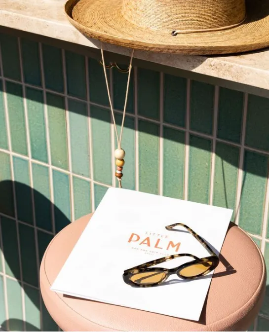 Straw hat, tortoiseshell sunglasses, and a Little Palm card on a pink poolside stool